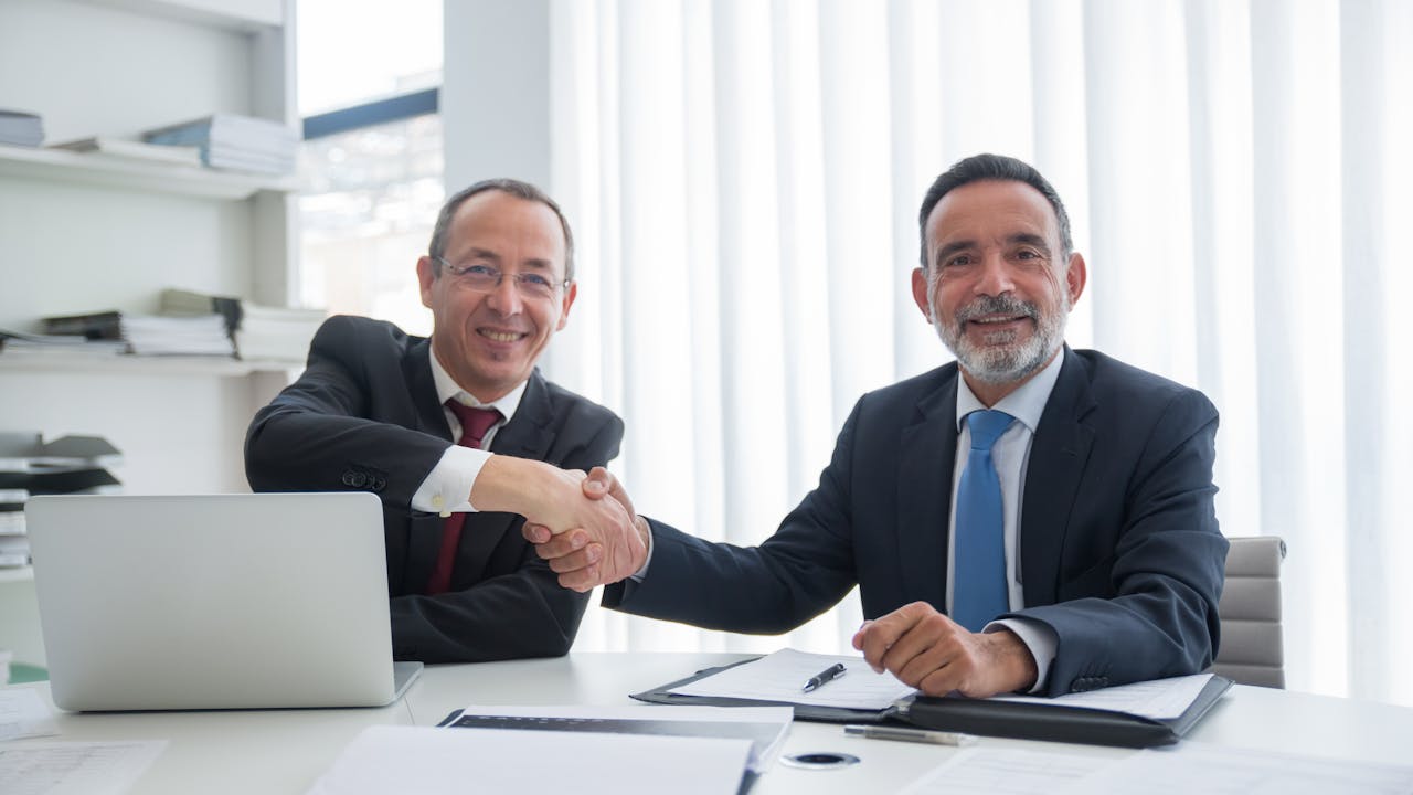 Two businessmen shaking hands at a desk in a professional office